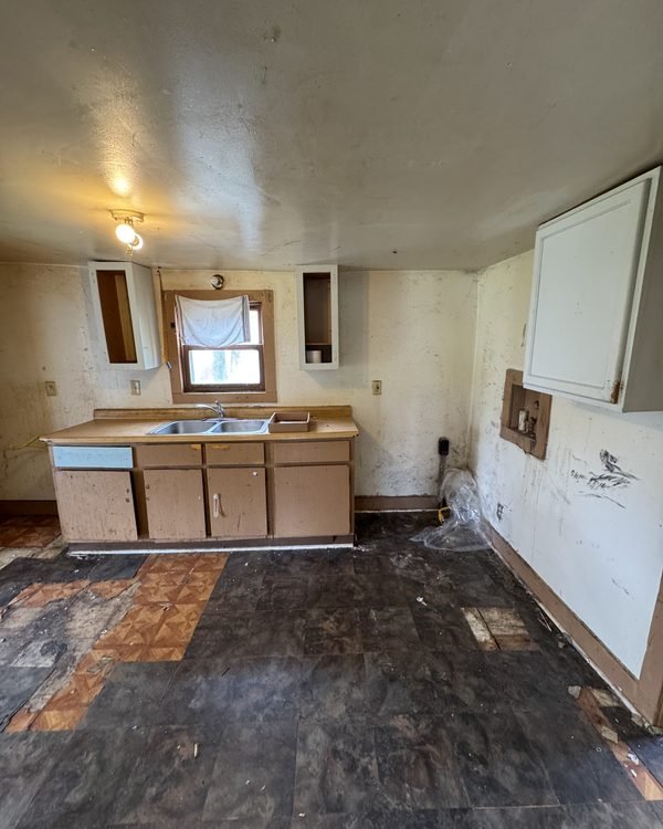 Kitchen with cabinets removed, exposed subfloor and stained ceiling, as found