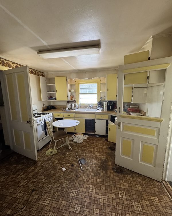 Dated kitchen with yellow cabinets, worn tile floor and items on the counters, as found